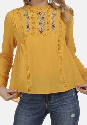 Femme portant un chemisier jaune moutarde avec broderies florales et panneaux en dentelle, associé à un jean bleu déchiré.