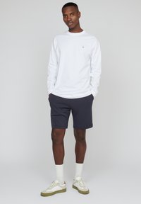 Balta ilgomis rankovėmis marškiniai, tamsiai mėlyni šortai ir pilki sportbačiai su žaliomis detalėmis. Modelis stovi su rankomis kišenėse, demonstruodamas atsipalaidavusį įvaizdį.