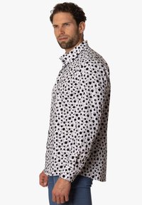 Camisa blanca con patrones circulares en negro y gris, de manga larga, con cuello abotonado y dobladillo recto. La tela parece suave y ligera.