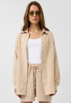 Kobieta w beżowej, teksturowanej koszuli zapinanej na guziki i pasujących szortach, z białym topem i zielonymi okularami przeciwsłonecznymi, stojąca na jednolitym tle.