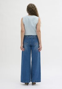 En ljusblå crop top i tyg, parat med vida blå jeans med en blekt textur och bakfickor. Modellens händer vilar vid sidorna.