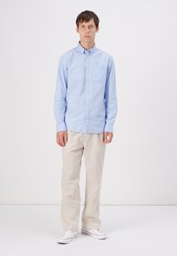 Light blue button-up shirt with a chest pocket, paired with beige pants and white sneakers. Simple design, woven texture, and fitted cut.