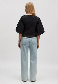Vrouw met lang blond haar, gekleed in een zwart top met pofmouwen en lichtblauwe wijde jeans, staande met haar gezicht naar weg van een effen achtergrond.