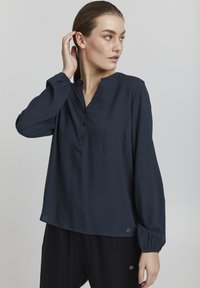 Blusa de manga larga en color navy, confeccionada en tela ligera, con escote en V, tapeta de botones y puños fruncidos, realzada con detalles metálicos en plata.