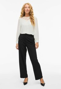 Weiße Bluse mit Spitzenverzierung am Ausschnitt, langen Ärmeln, kombiniert mit hochtaillierten schwarzen weiten Hosen und schwarzen spitzen Pumps.