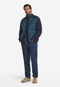 Vadderad teal väst med horisontell sömnad, bärs över en mörk skjorta. Blå jeans och ljusbruna skor fulländar outfiten.