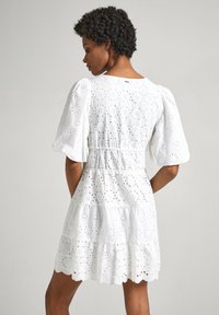 Robe blanche en broderie anglaise avec manches bouffantes, design à niveaux, motifs floraux et ourlet scallopé. Comprend une taille cintrée et un tissu texturé.