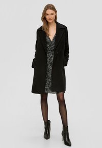 Schwarzer Wollmischmantel mit Reverskragen, seitlichen Taschen und geradem Schnitt, über einem gemusterten schwarz-weißen Kleid. Schwarze Ankle Boots.