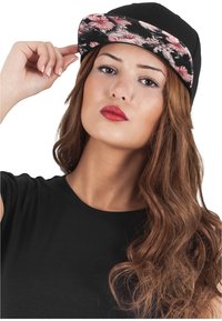 Fekete snapback sapka virágmintás peremmel. A modellen hosszú, hullámos barna haja van. Fekete pólót visel, és merész ajakrúzsot.