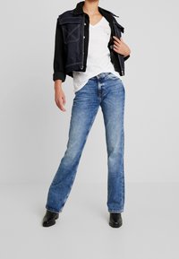 Chaqueta de mezclilla en azul oscuro con un diseño recortado, llevada sobre una camiseta blanca de cuello en V. Jeans rectos de color azul claro y botas de tobillo negras.