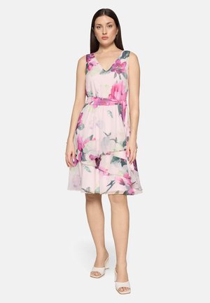 Blomstret kjole i pastelrosa med v-hals og ærmeløs design. Har en bælte i taljen og lagdelt, flæset kantdetalje. Båret med hvide sko.