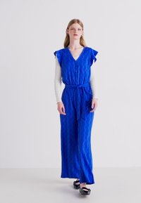 Blauwe, gestructureerde jumpsuit met korte, gerimpelde mouwen, een V-hals, knopen aan de voorkant, een gestrikte taille en wijde benen. Zwarte ballerina's maken de look af.