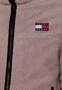 Beige fleecejakke med sort lynlås, med et rektangulært logoemblem i marineblå, hvid og rød på øverste venstre side. Tekstureret stof.