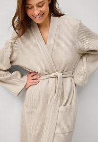 Beige wafelbreigoed met lange mouwen, een strik taille en twee voorzakken. Zachte textuur met een ruitpatroon, geschikt voor casual dragen.