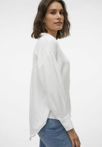 White, textured long-sleeve top with a relaxed fit and buttoned cuffs, paired with denim jeans, viewed from the side.