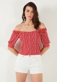 Red, off-the-shoulder blouse with puff sleeves, featuring a floral pattern. Worn with white denim shorts, highlighting a fitted design.