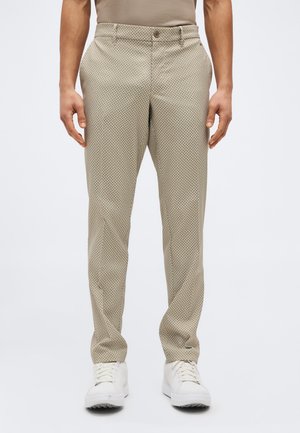 Μπεζ παντελόνι με σχέδιο ψαροκόκκαλο, φορεμένο με λευκά αθλητικά παπούτσια, εμφανίζεται από τη μέση μέχρι τα πόδια πάνω σε απλό ανοιχτόχρωμο φόντο.