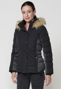 Crna i siva puffer jakna s kapuljačom obloženom krznom, horizontalnim i dijagonalnim šavovima, kopčama na dugme i bočnim džepovima.