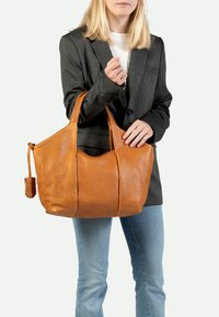 Bolso tote de cuero marrón con asas dobles, cuerpo grande y detalles de costura sutil, llevado por una persona que viste una chaqueta gris y jeans.