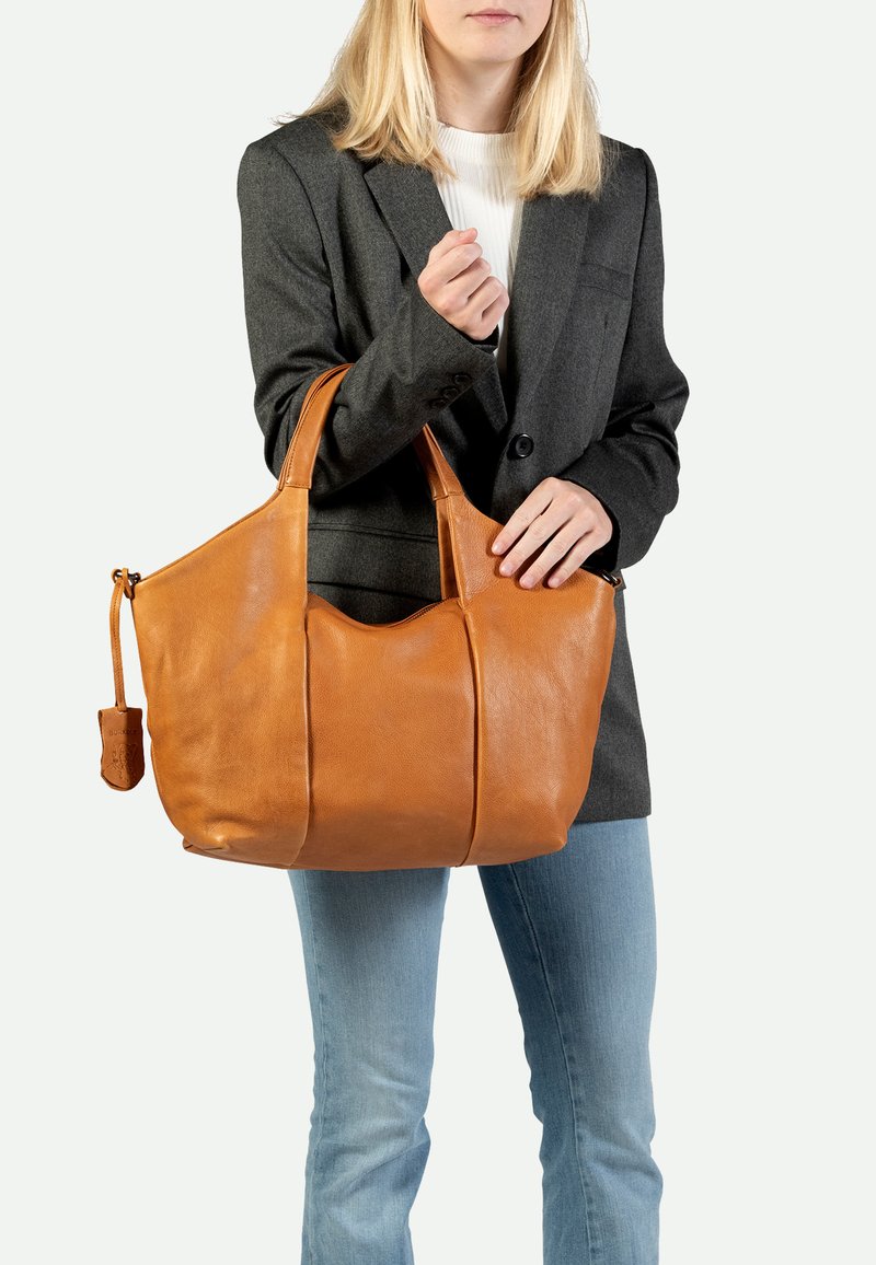 Bolso tote de cuero marrón con asas dobles, cuerpo grande y detalles de costura sutil, llevado por una persona que viste una chaqueta gris y jeans.
