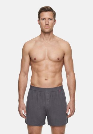 Fit, shirtloze man met kort haar, draagt grijze boxershorts en staat voor een effen witte achtergrond.