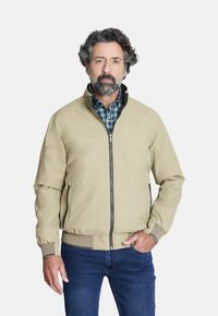 Chaqueta bomber beige con puños y dobladillo acanalados, cierre de cremallera, bolsillos laterales, superpuesta sobre una camisa de cuadros verde. Combinada con jeans azules.