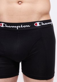 Ανδρική μέση που φοράει μαύρα μπόξερ με το λογότυπο "Champion" στη ζώνη, σε απλό φωτεινό φόντο.