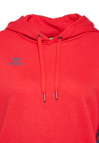 Felpa rossa in cotone, con tasca a marsupio frontale, cordini e un piccolo logo blu sul petto. Texture liscia.