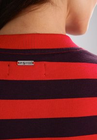 Suéter a rayas rojas y marinas con cuello acanalado. Presenta una pequeña etiqueta metálica en la parte posterior del cuello, con detalles de costura visibles.