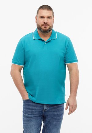 Mann mit Bart, der ein türkisfarbenes kurzärmeliges Poloshirt und blaue Jeans trägt, steht mit einer Hand in der Tasche vor weißem Hintergrund.