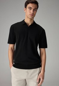 Sort kortærmet polo skjorte med V-hals, lavet af blødt stof. Har ribbet kant og krave, båret med lysebeige bukser.