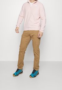 Felpa zip-up rosa chiaro, pantaloni cargo beige e scarpe da ginnastica blu con dettagli neri e gialli. Abbigliamento casual per attività all'aperto.