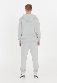 Man som står vänd bort ifrån kameran, iförd en grå hoodie och matchande sweatpants med vita sneakers, på en vit bakgrund.