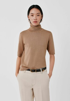 Femme aux cheveux noirs portant un pull beige à col roulé manches courtes, un pantalon beige clair, une ceinture noire et des boucles d'oreilles créoles dorées sur fond blanc.