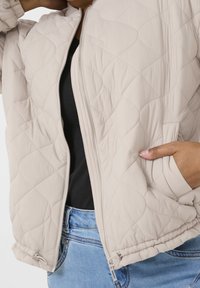 Person klädd i en beige vadderad zip-up jacka över en svart topp och blåjeans, med en hand i jackfickan.