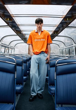 Jonge man in fel oranje shirt en lichtblauwe spijkerbroek staat in een lege bus met blauwe stoelen en glazen dak op een regenachtige dag.