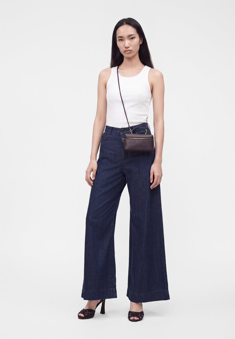Femme portant un débardeur blanc, un jean bleu foncé à jambes larges, des talons noirs ouverts et un petit sac bandoulière marron, debout devant un fond uni.
