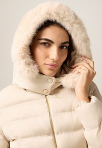 Beżowa kurtka puchowa z kapturem obszytym sztucznym futrem, wyposażona w złoty zamek błyskawiczny i zatrzaski, z pikowaną teksturą i dopasowanymi mankietami.