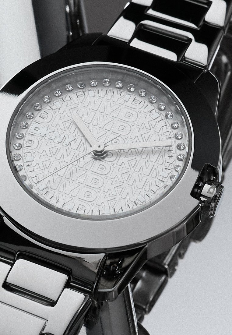 Reloj metálico plateado con un dial blanco texturizado elevado que presenta un patrón y acentos de cristal. Diseño de pulsera de eslabones de metal cepillado.