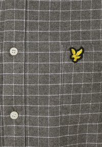 Grå flanellskjorta med ett vitt rutmönster, med en gul broderad fågel-logotyp på vänster sida och vita knappstängningar.