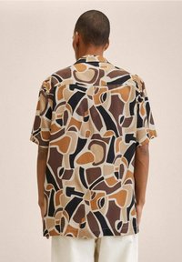 Mann mit einem kurzärmeligen Hemd mit einem abstrakten geometrischen Muster in Beige, Braun, Schwarz und Weiß, der mit dem Rücken zu uns steht.