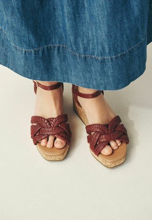 Fødder iført brune flettede læderplatformsandaler med ankelremme, delvist dækket af et langt blåt denimskørt.