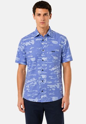 Jeune homme portant une chemise bleue à manches courtes avec des motifs abstraits bleu clair et le logo « BOSS » sur la poitrine, associée à un pantalon sombre.