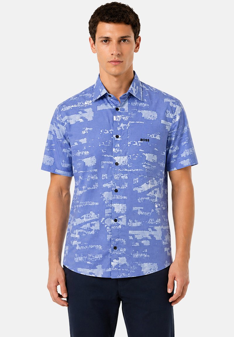 Jeune homme portant une chemise bleue à manches courtes avec des motifs abstraits bleu clair et le logo « BOSS » sur la poitrine, associée à un pantalon sombre.