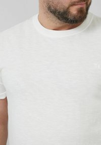White short-sleeve t-shirt made of a soft, textured fabric. Features a round neckline and minimal branding on the side.