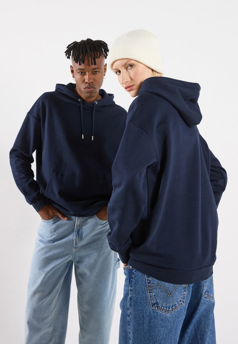 Marine hettejakker med snøring, avslappet passform, med kengurulommer. Én modell har på seg en kremfarget lue; begge har lyse denimjeans.