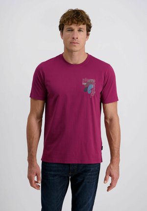 Mann in einem kurzärmeligen magentafarbenen T-Shirt mit "Pierre Cardin"-Logo auf der Brust, kombiniert mit dunkelblauen Jeans, vor einem schlichten Hintergrund.