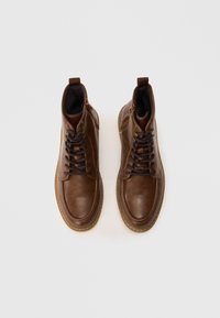 Braune Leder-Stiefeletten mit einer glatten Textur, ausgestattet mit Schnürsenkeln, seitlichen Reißverschlüssen und einer Gummisohle. Mit gesteppten Akzenten durchgehend.