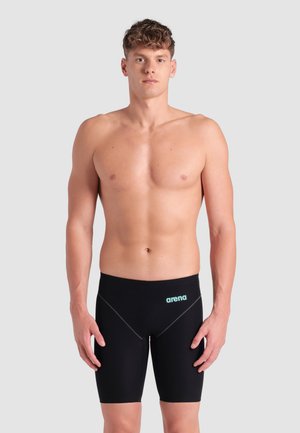Mladý muž s krátkymi kučeravými vlasmi, oblečený v čiernych plaveckých šortkách značky Arena, stojaci na jednoduchom svetlosivom pozadí.
