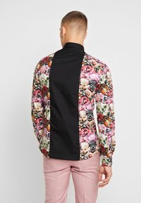 Chemise à manches longues florale avec panneau noir, présentant des motifs floraux multicolores et une texture lisse. La vue de dos montre un design distinct.